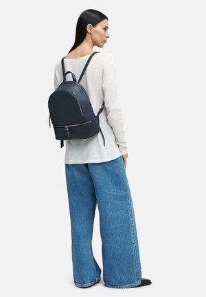 Donna con capelli lunghi e scuri che indossa una maglia bianca a maniche lunghe, jeans blu a gamba larga, scarpe nere con punta e uno zaino di pelle scura.