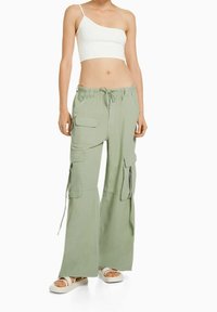 Ljusgröna cargobyxor med dragsko i midjan, flera fickor och siddragkedjor, tillsammans med en vit crop top med en axel och sandaler.