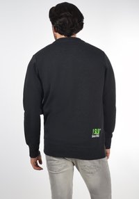 Black sweatshirt with a textured finish, crew neck, and ribbed cuffs. Green logo with text on the lower back. Gray jeans visible below.