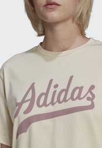 Grietai spalvos medvilnės marškinėliai su dideliu rožiniu "Adidas" logotipu, apvaliu iškirpimu, trumpomis rankovėmis ir laisvu siluetu.