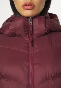 Close-up van de lippen en kin van een persoon die een gewatteerde bordeauxrode jas met rits en hoge kraag draagt.