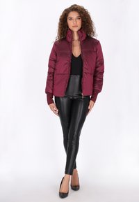 Burgundi színű pufferszűkítő dzseki magas gallérral és zipzárral, fekete műbőr nadrággal és szegecselt fekete magassarkú cipővel párosítva. Az anyag sima textúrájú.