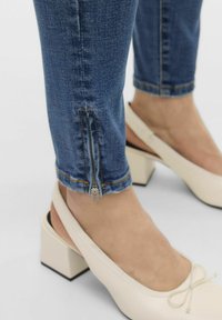 Jean en denim bleu avec un détail de zip à la cheville, associés à des chaussures à talons blocs crème ornées d'un noeud. Texture lisse et coupe classique.