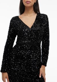 Sieviete, tērpusies garām piedurknēm, melnā sequined V veida kakla kleitā, attēlota no pleciem līdz gurniem uz balta fona.