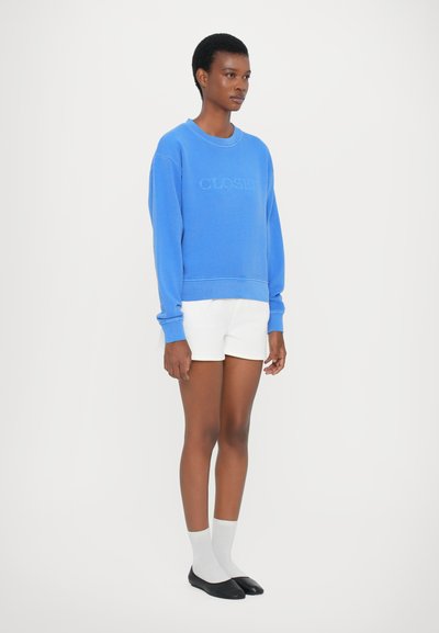 Μπλε φούτερ με υφή στο λογότυπο "CLOSER", συνδυασμένο με λευκά σορτσάκια. Το μοντέλο φορά γκρι κάλτσες και μαύρες μπαλαρίνα, ενώ στέκεται σε λευκό φόντο.