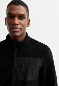 Svart fleecejacka med hög krage, som har ett texturerat, mjukt tyg och en slät nylonframsida. Dragkedjestängning och minimalistisk design.