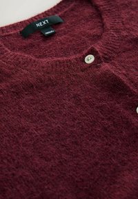 Bordeaux strikket sweater med rund hals, der har en tekstureret overflade, to hvide knapper og et mærkeskilt ved kraven.