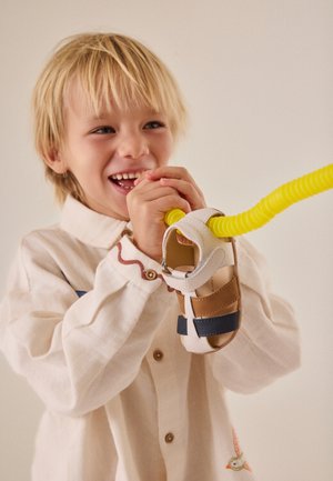 Niño rubio sonriente sosteniendo una sandalia de niño pequeño sujeta a una cuerda amarilla en espiral sobre un fondo neutro.