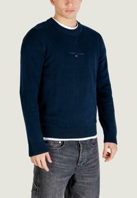 Maglione blu navy lavorato a maglia con polsini e orlo a costine, caratterizzato da un piccolo logo ricamato al centro. Indossato sopra una camicia bianca e jeans scuri.