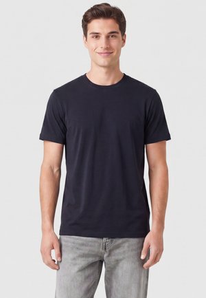 Junger Mann trägt ein schlichtes schwarzes T-Shirt mit kurzen Ärmeln und hellgraue Jeans, steht lächelnd vor einem einfarbigen Hintergrund.