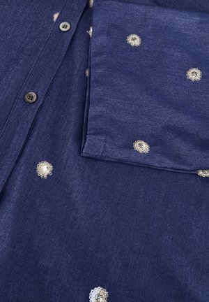 Blauw denimshirt met kleine witte geborduurde bloemen en zichtbare knoopsluiting, met een deel van de lange mouw en knopdetails zichtbaar.