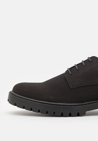 Chaussure en suède noir avec un bout rond, un design à lacets plats et une semelle en caoutchouc épaisse avec une texture nervurée. Accents de coutures minimales.