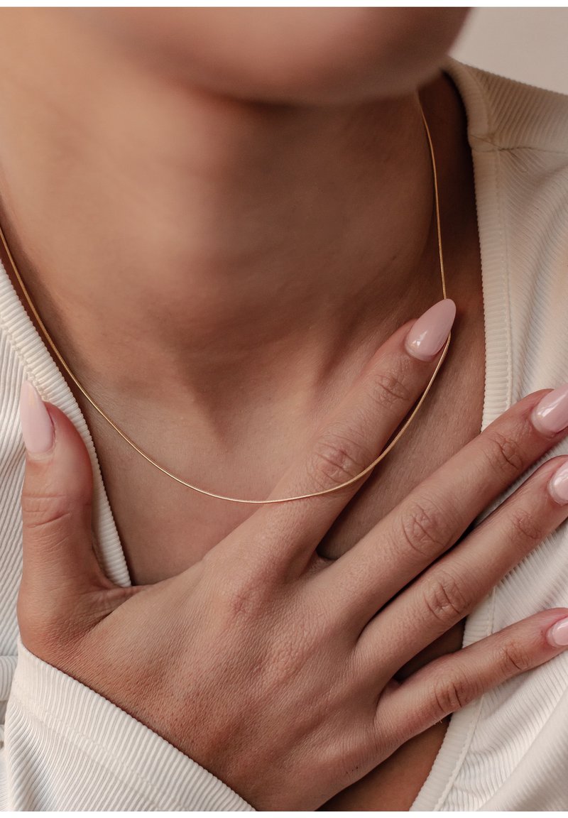 Collier en chaîne dorée reposant sur la peau, avec un design lisse et fin. La main du modèle, avec des ongles rose clair, effleure légèrement le collier.