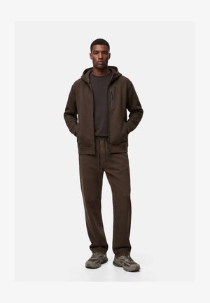 Junger Mann steht, trägt eine braune Kapuzenjacke mit Reißverschluss, passende braune Hose, dunkelbraunes Hemd und graue Sneakers, Hände in den Taschen.