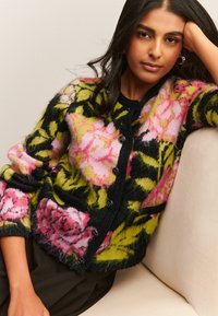 Femme aux cheveux bruns portant un cardigan pelucheux à motifs floraux dans des couleurs rose, vert et noir, assise sur un canapé beige clair.