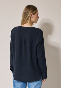 Camicia a maniche lunghe di un blu navy scuro realizzata in tessuto testurizzato, con una piega sul retro, orlo arrotondato e polsini con bottoni. Indossata con jeans azzurri.