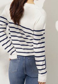 Maglione bianco a righe con linee orizzontali blu navy, dotato di scollo a girocollo e polsini a costine, abbinato a jeans blu aderenti.