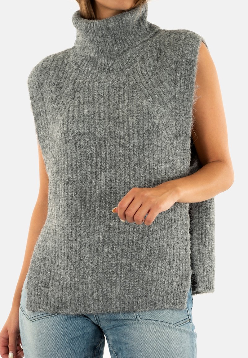 Donna che indossa un maglione a collo alto grigio senza maniche e jeans azzurri chiari, accorciati sulle spalle e sui fianchi.