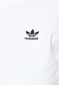 Balta kokvilnas T-kreklu ar melnu izšūtu Adidas logotipu trīs lapu dizainā. Audumam ir gluda, maiga tekstūra.