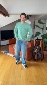 Jeune homme en sweat-shirt vert et jean bleu debout dans un salon avec un sol en bois, un canapé orange, une grande plante et du matériel d'exercice.