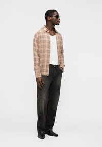 Mann mit Sonnenbrille, beigem kariertem Hemd über weißem Oberteil, schwarzen, ausgewaschenen Jeans und schwarzen, spitzen Schuhen, stehend mit einer Hand in der Tasche.