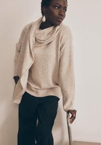 Maglione oversize beige con colletto drappeggiato, polsini a costine e orlo grezzo, abbinato a pantaloni neri. Tessuto morbido e vestibilità comoda.