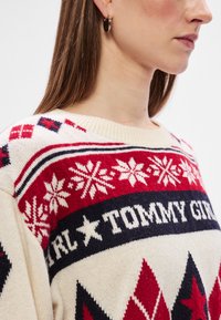 Krém színű pulóver piros, fehér és sötétkék mintázattal, beleértve a hópelyheket és a gyémántokat. Kerek nyakkivágással és kötött textúrával rendelkezik.