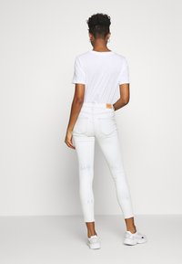 Vaqueros ajustados de tiro alto blancos con acentos de tie-dye azul claro, con una etiqueta en la parte posterior, combinados con una camiseta blanca sencilla y zapatillas.