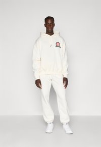 Κρεμ φούτερ με μπροστινή τσέπη και πράσινο κεντητό λογότυπο, σε συνδυασμό με συμβατά παντελόνια φόρμας. Φοράει λευκά αθλητικά παπούτσια.