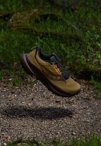 Pantof de alergare trail maro și negru suspendat deasupra unei cărări cu pietriș, cu iarbă verde și vegetație în fundal.