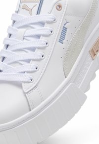 White leather sneaker with textured side accents, chunky white sole, and flat laces. Features a logo on the tongue and side.