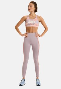 Crop top sportivo in rosa pastello con logo bianco e motivo foglia; abbinato a leggings a vita alta lilla chiari con strisce laterali arancioni.