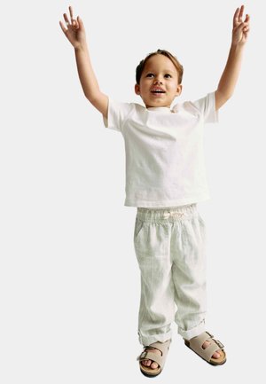 Witte korte mouwen t-shirt, lichtgrijze gebloemde broek met elastische taille, en bruine sandalen met twee gespen, met opgestoken armen.
