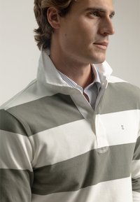 Polo a rayas en verde oliva y blanco, hecho de algodón. Cuenta con un collar, una tapeta de botones y un logo bordado en el pecho.