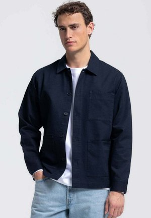 Jonge man die een marineblauw jasje met knopen draagt over een wit t-shirt en lichtblauwe spijkerbroek, staat met één hand in de zak.
