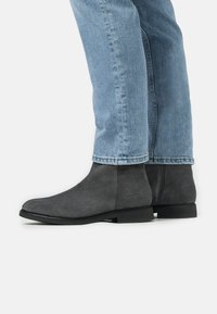 Bottines en daim gris avec une pointe arrondie élégante, une semelle noire plate et un design minimaliste. Présente un logo de marque subtil sur le côté.