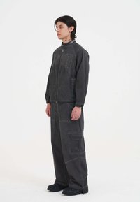 Giacca grigia, strutturata, con zip, caratterizzata da dettagli ricamati, abbinata a pantaloni cargo coordinati. Calzature nere e robuste completano il look.