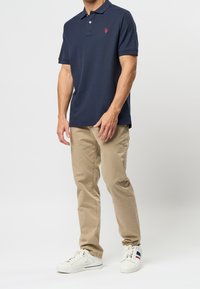 Granatowa koszulka polo z kołnierzykiem i czerwonym logo, w zestawieniu z beżowymi spodniami i białymi trampkami. Gładka tkanina o dopasowanym kroju.