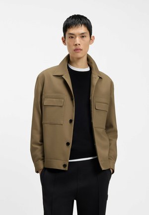 Ung mand iført en beige jakke med knapper og store lommer over en sort sweater og sorte bukser, stående foran en ensfarvet hvid baggrund.
