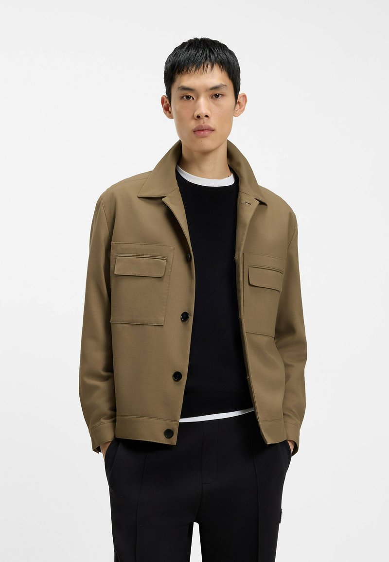 Jeune homme portant une veste beige boutonnée avec de grandes poches par-dessus un pull noir et un pantalon noir, debout devant un fond blanc uni.