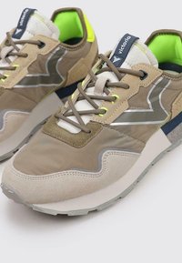 Chaussures de sport beiges et vertes en matériaux en cuir suédé et textile, avec des accents réfléchissants et une semelle blanche épaisse à motif texturé.