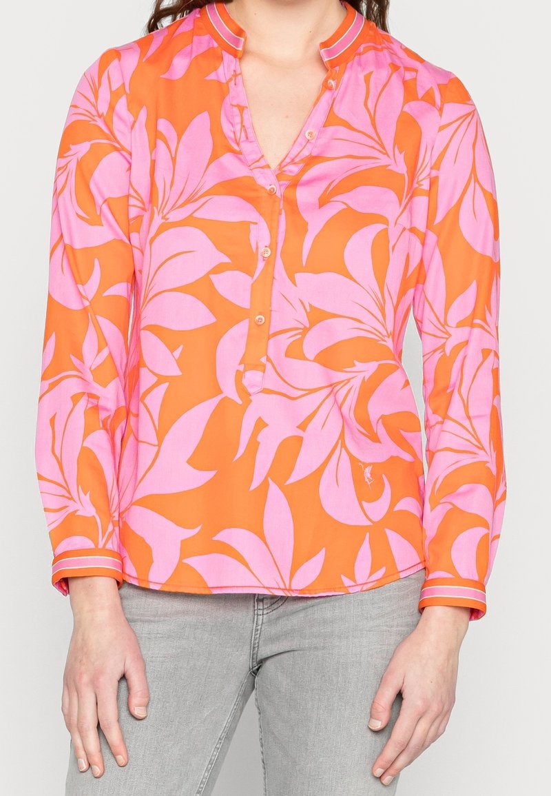 Femme portant un chemisier orange à manches longues avec de grands motifs floraux roses et un pantalon gris clair, debout, les mains détendues le long du corps.