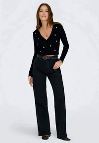 Črn skrajšan kardigan z motivi srca, v kombinaciji z visokimi črnimi hlačami in črnimi čevlji z zajetim prstom, okrašenimi s studenci.