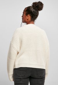 Krämfärgad stickad tröja med ribbad textur, vida ärmar och croppad passform, matchad med mörka jeans. Bakvyn visas.