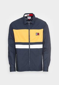 Marinblå zip-up jacka med gul horisontell rand, vit accent och logotypapplikation. Tillverkad av bomull, har en krage och långa ärmar.