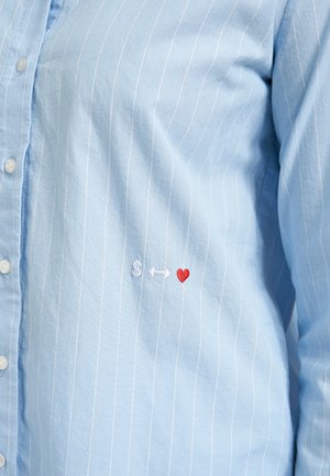 Light blue pinstriped shirt with small embroidered white dollar sign, white double-headed arrow, and red heart near the waist.