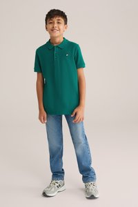 Zöld póló ing gallérral, három gombbal és egy kis logóval, világos kék farmernadrággal és szürke sportcipővel párosítva.