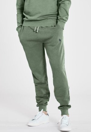 Groene katoenen sweatpants met elastische tailleband en aansluitende enkels, voorzien van zijzakken en een klein marineblauw logo. Gecombineerd met witte sneakers.