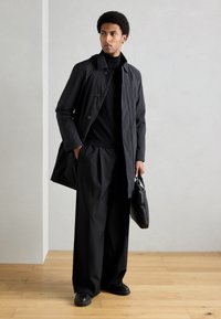 Fekete ballonkabát gombos záródással, fekete garbó és bő szárú fekete nadrág fölött viselve. A modell fekete táskát tart.