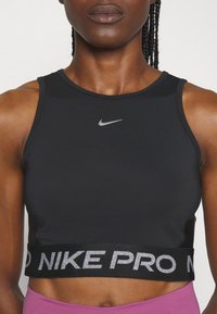 Femme athlétique portant un crop top noir sans manches avec un logo Nike argenté et une large ceinture de marque, associée à un legging rose.